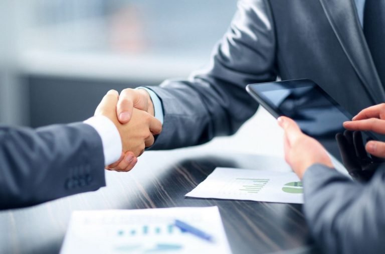 Picture of two people in business suits shaking hands over papers laying on a table.